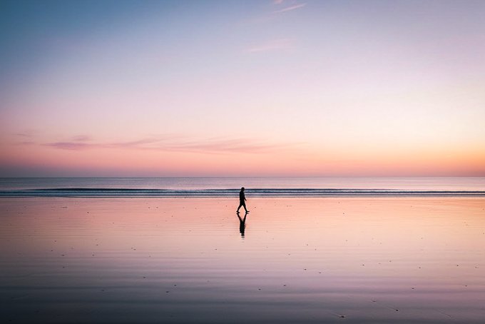 A serene photograph of a lone person walking on a wet beach at sunset, with a gradient sky of pink and blue reflected perfectly on the smooth sand.