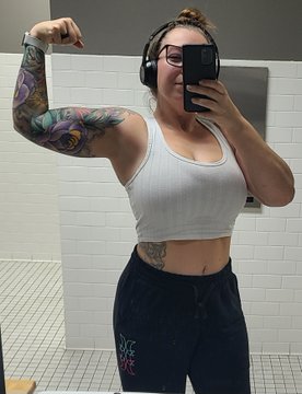 A woman with a tattooed arm and midriff tattoos takes a mirror selfie in a bathroom wearing a white crop top, headphones, and dark sweatpants while flexing her bicep.