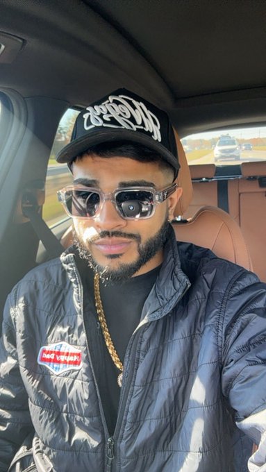 A young man with a beard, wearing sunglasses, a black cap with white script lettering, and a dark jacket with a red patch, takes a selfie inside a car.
