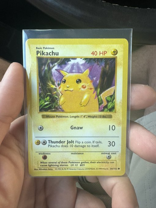 A hand holding a first edition Pikachu Pokémon trading card in a protective sleeve, showing 40 HP with attacks Gnaw and Thunder Jolt.