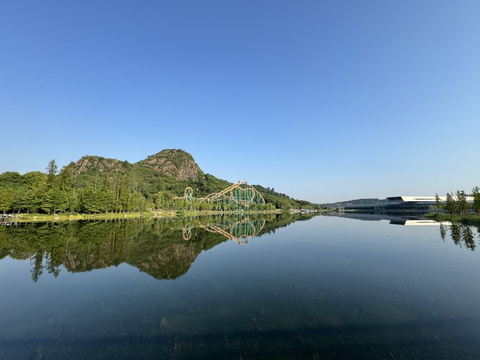A serene lake reflects a mountain, lush greenery, and a roller coaster under a clear blue sky, captured during the poster's morning run in Suzhou, where they enjoyed a scenic anaerobic training session with #STEPN.