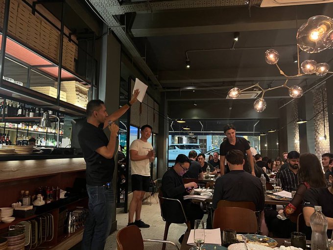 A man in a black shirt speaks into a microphone while holding up a paper, addressing a group of diners seated at tables in a dimly lit, modern restaurant. The scene captures a social gathering that aligns with the post's reference to an OML-related event at a Sentient AGI dinner, with attendees engaged in conversation and dining.