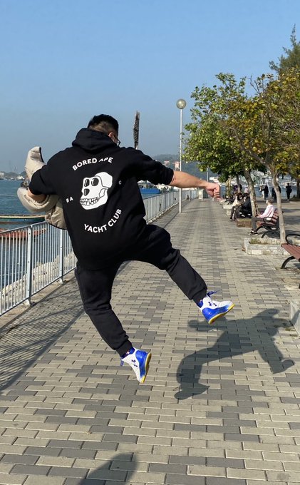 A person leaps joyfully on a waterfront promenade wearing a black "BORED APE YACHT CLUB" hoodie featuring the iconic skull logo, capturing the celebratory spirit of being part of the renowned NFT community.