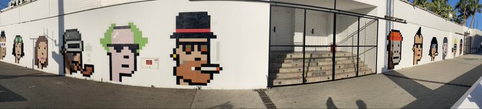 A panoramic street view shows a building's white exterior wall decorated with pixel-art style murals of various characters, including one with green hair and another with a top hat, next to a gated entrance with stairs.