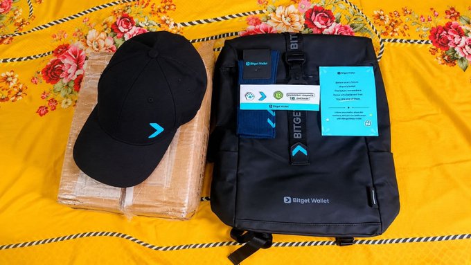 A black backpack with 'Bitget Wallet' branding sits alongside a black cap, both featuring teal Bitget logos, displayed on a floral-patterned yellow surface.