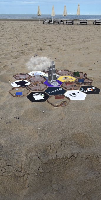 An aerial view of a hexagonal game board called 'Tik Tak Battle' arranged on a sandy beach, featuring cartoon animal characters and a smoking central tower, with closed umbrellas and lounge chairs in the background.