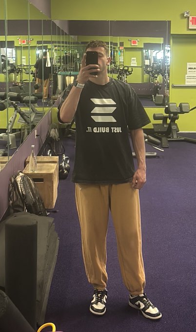 A man takes a mirror selfie in a gym, wearing a black t-shirt with a white logo and 'Just Build It.' text, paired with tan sweatpants and black and white sneakers.