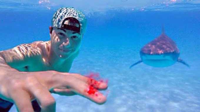 An underwater selfie showing a smiling man wearing a backwards cap with a bleeding finger, while a shark approaches in the background.