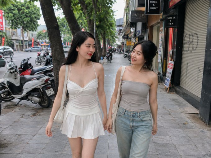 Two young women walk hand-in-hand down a tree-lined urban sidewalk, the one on the left in a white mini dress and the other in a gray tube top and light-wash jeans, with storefront signs like "BOLD & LIGHT," "PATEV," and "COFFEE & TEA" visible behind them.

The image's depiction of two people connected in a real, visible bond walking through a neighborhood of local shops visually echoes the post's message about authentic, proximity-based connections—reinforcing that genuine influence, like genuine friendship, comes from real closeness rather than manufactured metrics.