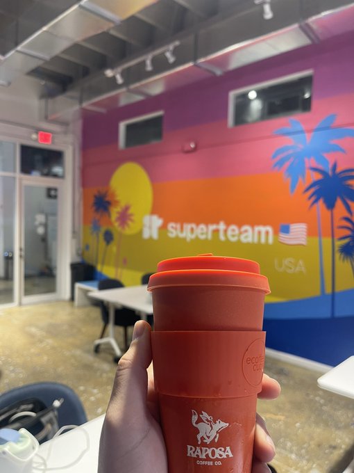 A hand holds an orange Raposa Coffee Co. reusable cup in front of a colorful 'Superteam USA' mural featuring palm trees and a sunset design.