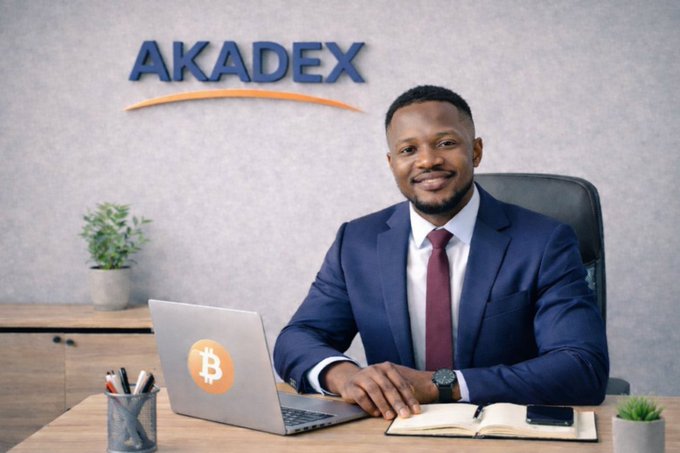 A smiling man in a blue suit and burgundy tie sits at a desk with a Bitcoin-logo laptop in front of an 'AKADEX' wall sign.