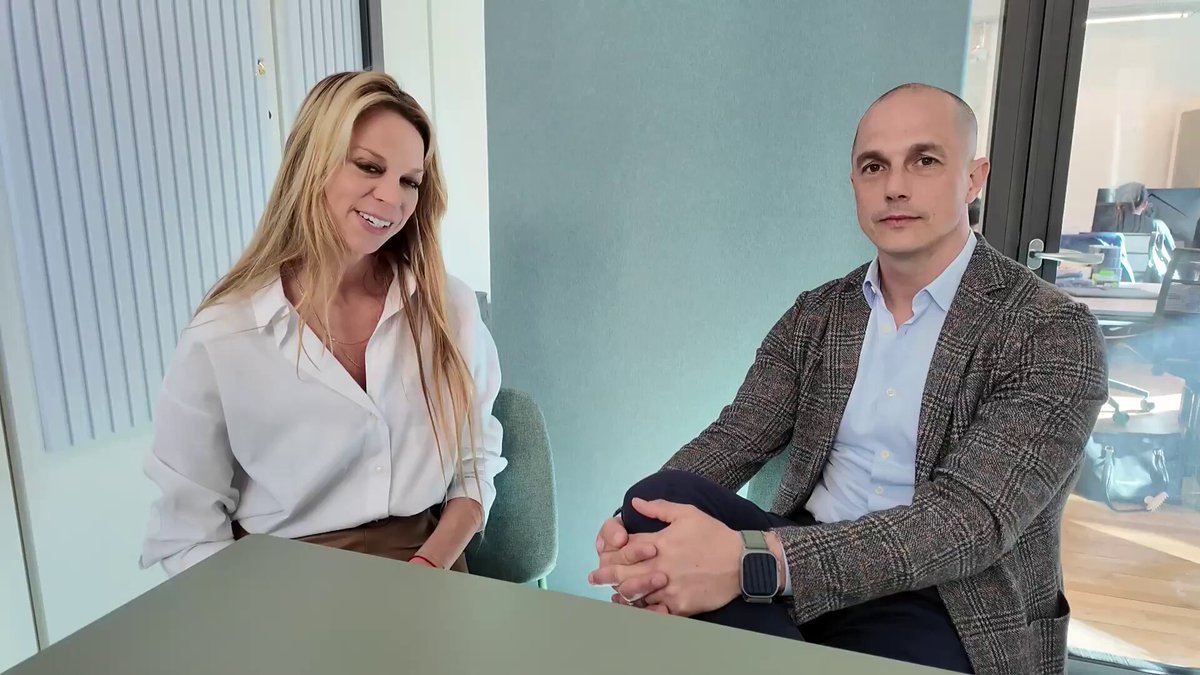 The image shows a man and woman sitting at a desk, with their hands clasped together. They are both wearing business attire, suggesting they might be professionals or attending a formal event. The background of the image reveals that they are in an office setting, which further supports the idea that this is a professional environment.