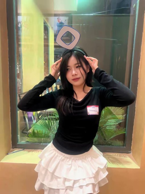 A young woman with long black hair stands by a window, wearing a black long-sleeve top with a name tag and a tiered white skirt, posing playfully with her hands near her head and a stylized "MON" logo floating above her—reflecting a fun moment from last year's Vietnam meetup, as the post nostalgically references her return with the ticker $MON.