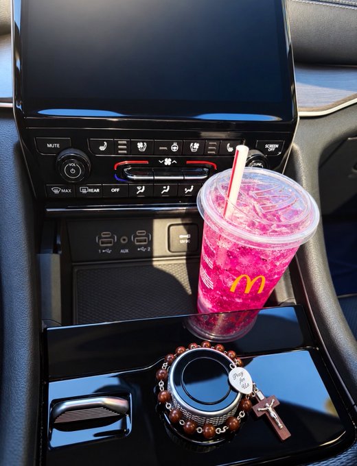 A car interior console holding a McDonald's cup filled with a pink drink, positioned above a round beaded bracelet with a cross charm resting on the cup holder.