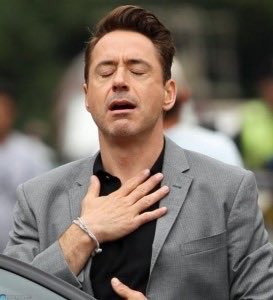 A man in a grey blazer and black shirt with his hand on his chest and eyes closed, appearing emotional.
