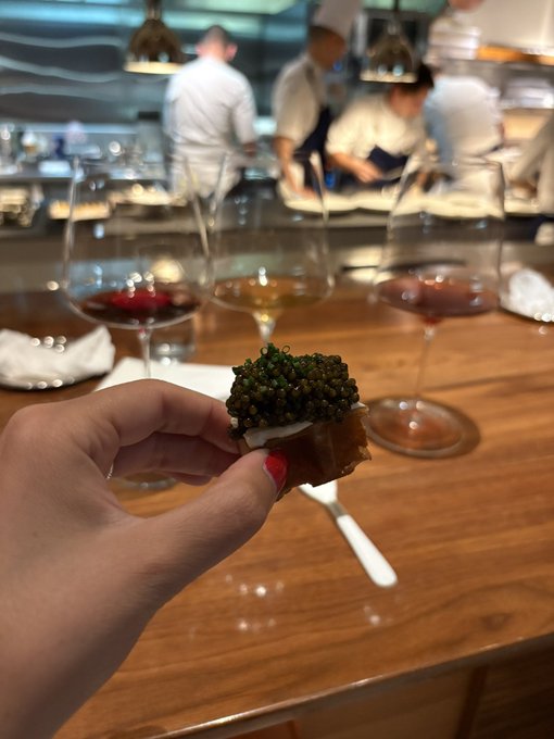 A hand holds up a small spoon of caviar at what appears to be an upscale restaurant's bar, with wine glasses and chefs visible in the background. The image complements the post's sentiment by showcasing an indulgent luxury moment worth savoring—the caviar becomes the focal point of the experience, representing those fleeting pleasures that matter more than material possessions.