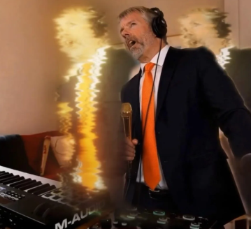 A man with a beard wearing a suit and orange tie sings into a microphone while standing behind an M-Audio keyboard in a room with warm lighting.