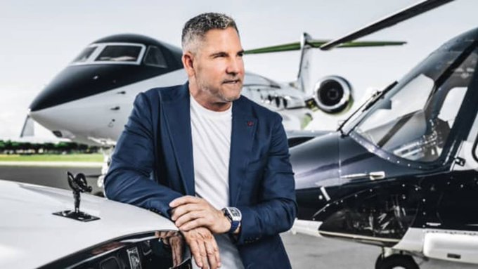 A man in a navy blazer and white t-shirt leaning on the hood of a black vehicle, with a private jet (registration N711) in the background.