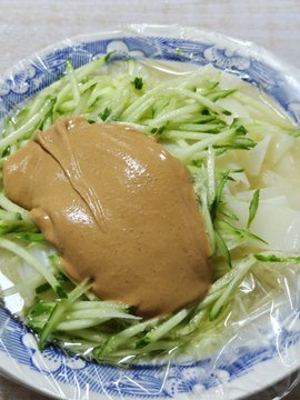 The image shows a bowl of cold skin noodles (凉皮) topped with shredded cucumber and a dollop of sesame paste, served in a blue and white patterned bowl. This simple, traditional meal matches the post's mention of enjoying an affordable 2.5U lunch at a decades-old shop, where the $BB stock profits help fund both the modest meal and Pokémon card collecting.