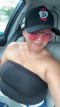 A woman wearing a black cap, pink sunglasses, and a black strapless top is sitting in a car, looking at the camera.