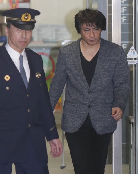 A serious-looking man with tousled dark hair, wearing a gray blazer over a black shirt, is escorted by a uniformed Japanese police officer with a peaked cap and gold badge insignia. The scene relates to reports of fans being defrauded by a cryptocurrency scheme associated with him, involving over 6,000 victims and approximately 100 million yen in losses.
