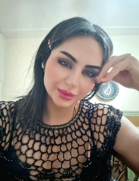 The image features a woman with long dark hair wearing a black top that has a mesh pattern. She is posing for a photo, looking directly at the camera while resting her hand on her face. The background of the image appears to be a blurred room or space filled with various objects and decorations.