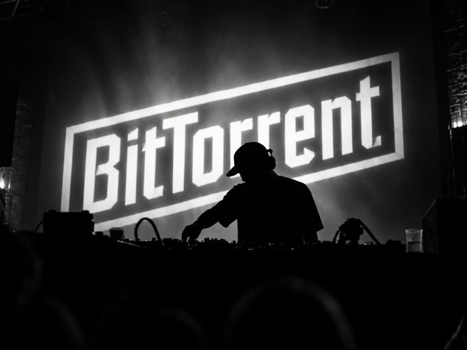 A silhouetted DJ stands behind a mixing deck in front of a large illuminated screen displaying the "BitTorrent" logo.