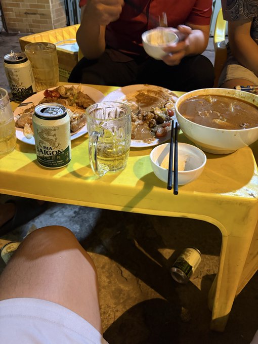 A casual dining scene at a low yellow plastic table showing leftover plates of food, a large bowl of soup, chopsticks, beer glasses, and a can of 'BIA SAIGON' beer, with people sitting around.