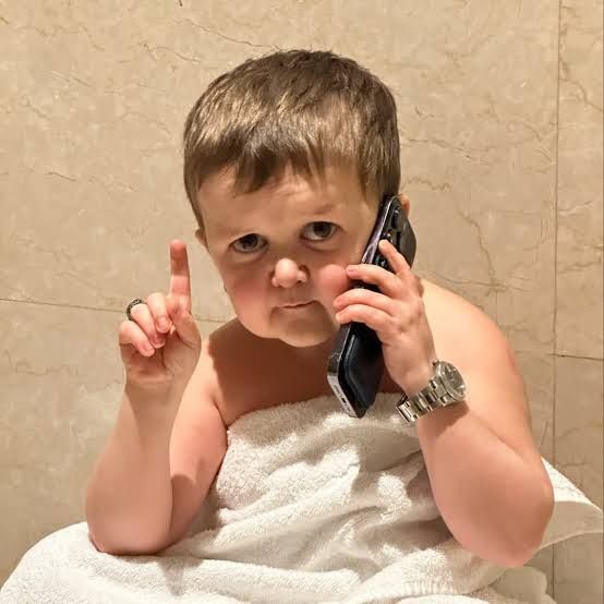 The image features a young boy sitting on a bathroom counter, wrapped in a towel while talking on his cell phone. The boy appears to be making a funny face as he engages in conversation. The background of the image shows a bathroom setting with a tiled wall and a sink visible.