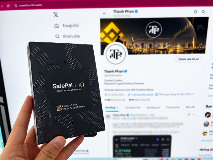 A hand holds a black SafePal X1 hardware crypto wallet in front of a computer screen displaying an X (formerly Twitter) profile for 'Thành Phan' (@thethaophan), described as a Content Creator for Research.Crypto/Andregg.Greenspy.