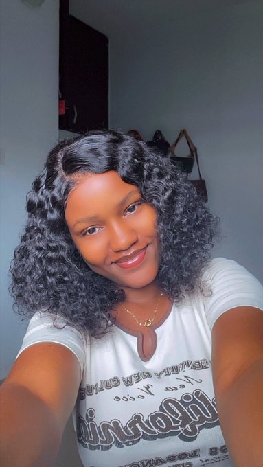 A selfie of a smiling young woman with curly black hair wearing a white t-shirt, standing in a room with a bag visible in the background.