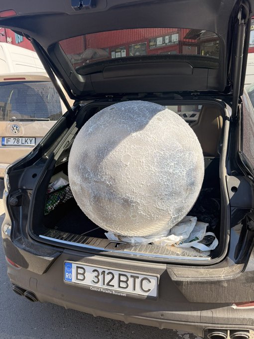 A large spherical object resembling the Moon is packed inside the open trunk of a black SUV with Romanian license plate 'B 312 BTC', parked next to a Volkswagen van with plate 'IF 78 LKY'.