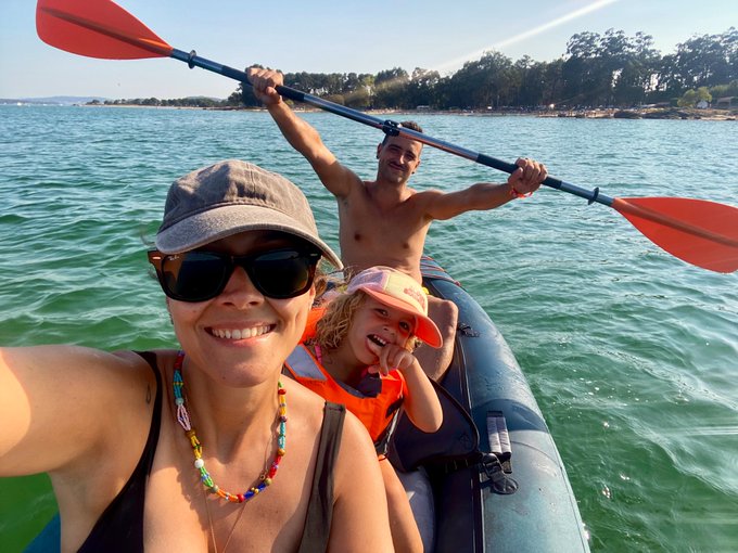 A family of three enjoys a sunny day kayaking together, with the woman smiling for a selfie while the man holds up the paddle and their young child laughs joyfully, perfectly capturing the warm, late-summer family moment described in the post.