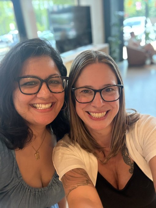 Selfie of two smiling women wearing glasses, one with dark hair and the other with lighter brown hair and arm tattoos, indoors.