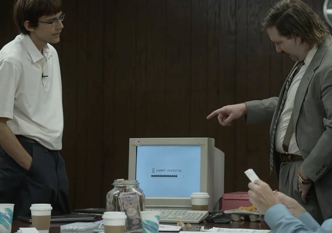 Two men in office attire stand at a desk looking at an old CRT monitor displaying "payment processing" with a loading bar, while coffee cups and snack bags clutter the foreground—evoking the grind of early tech startup life that the post nostalgically references with "Build on @arc."