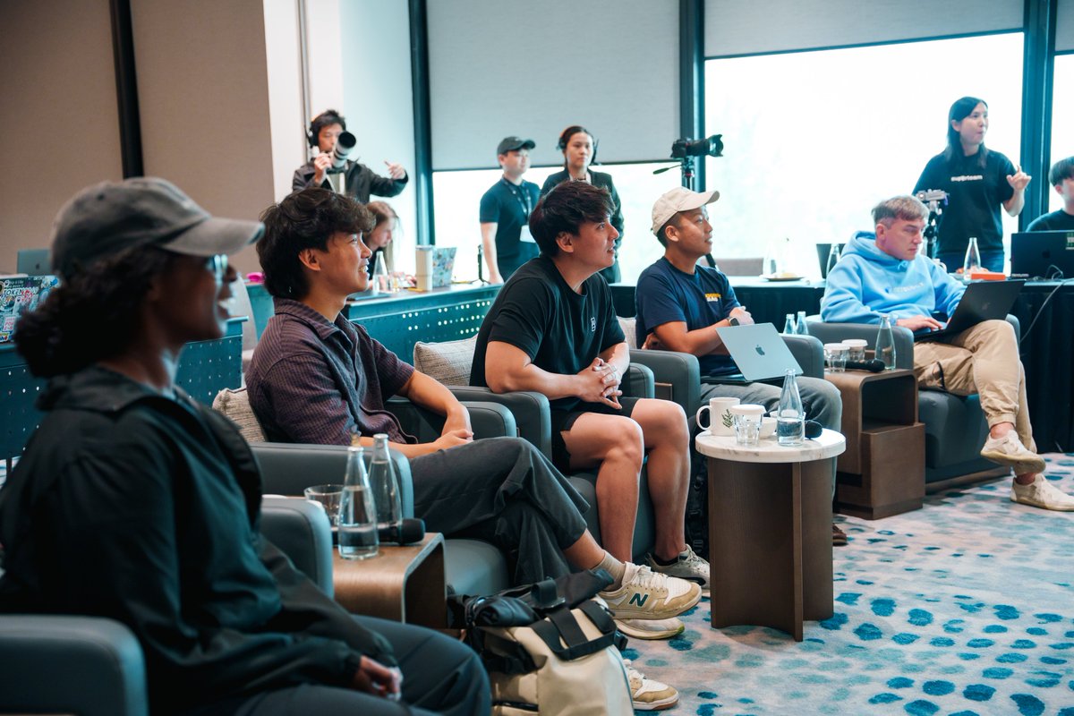 A group of casually dressed people sitting on chairs in a conference or event space, attentively watching a presentation with laptops on their laps and coffee mugs on side tables, while staff stand in the background.