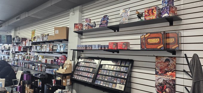 The interior of a trading card or collectibles shop, displaying shelves stocked with various game boxes, cards, and merchandise on white slatwall paneling.