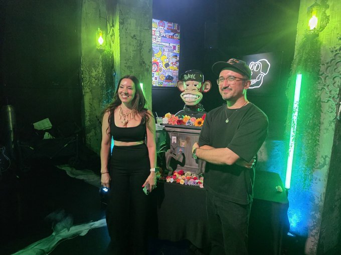 Two people stand smiling in a dimly lit venue with green neon lighting, posing next to a monkey sculpture wearing a cap with 'GANG' visible on it and a 'SKULLPANDA' neon sign in the background.