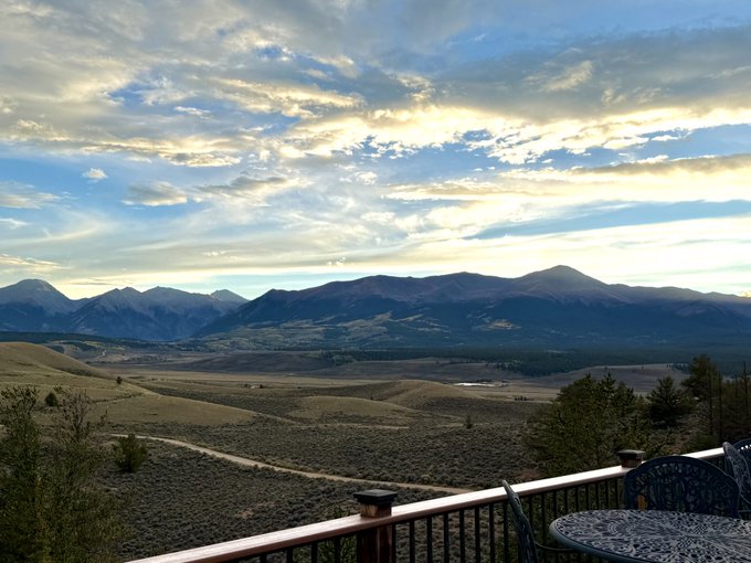 A scenic mountain landscape viewed from a deck with a railing and patio furniture in the foreground, showing rolling hills and a dramatic cloudy sky at sunset.