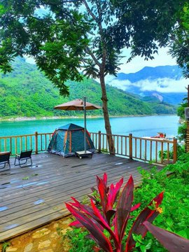 A serene lakeside view featuring a wooden deck with a green tent and umbrella, surrounded by lush green trees, red foliage, and mountains partially obscured by clouds in the background.