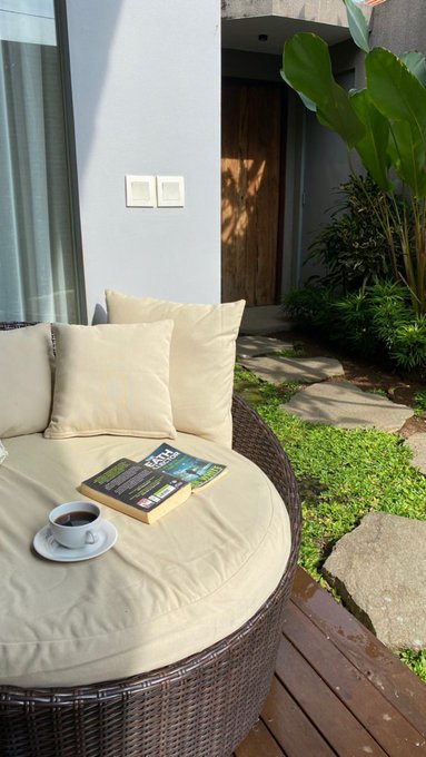 An outdoor patio scene featuring a round wicker daybed with beige cushions, holding a coffee cup and a book, set on a wooden deck beside a stone pathway and tropical plants.