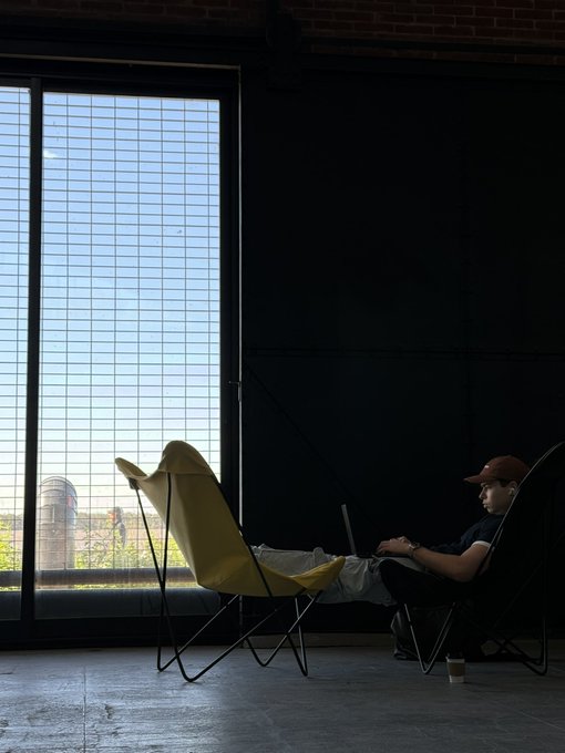 Esteban Suarez is seen working on his laptop in a modern space with large windows, possibly representing a focus on remote work or "tokenmaxxing."