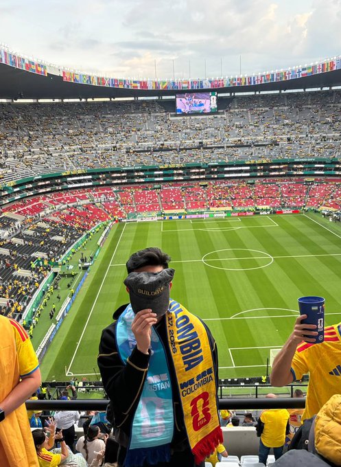 The image shows a large stadium filled with people, including fans wearing yellow shirts holding drinks such as cups of beer or soda. A man stands out from the crowd by holding a soccer scarf that says "Colombia" on it. The photo is taken from an elevated perspective, providing a bird's eye view of the scene and emphasizing the excitement and energy in the stadium. The post about the match day seems to be related to this event, as indicated by the man wearing a soccer scarf and the fans holding drinks.