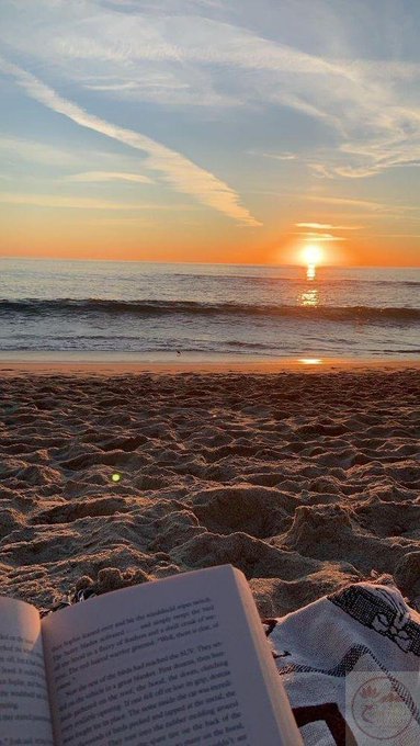 A scenic beach photo at sunset showing a person reading a book in the foreground with the ocean and sun setting on the horizon under wispy clouds.