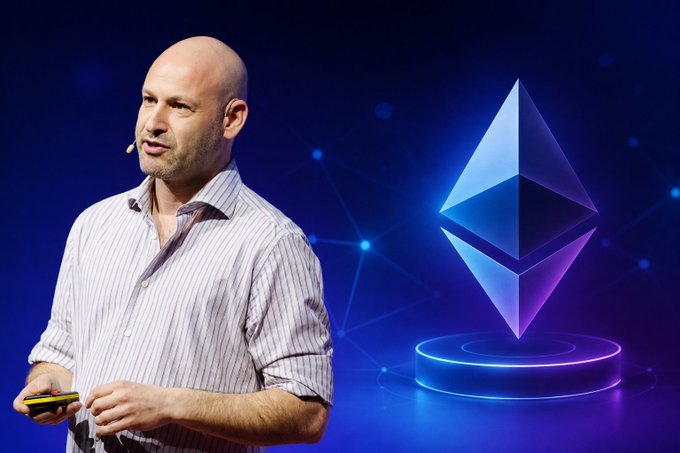 A bald man wearing a striped shirt and headset microphone speaks at a presentation, holding a clicker, with a glowing 3D Ethereum (ETH) logo displayed prominently on a blue digital background beside him. The image visually complements the accompanying post about Ethereum co-founder Joseph Lubin discussing the importance of low mainnet transaction fees for network adoption, staking, and ETH burning.