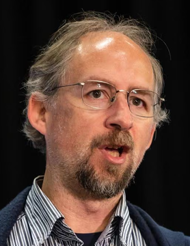 The image shows Adam Back, a prominent figure in the Bitcoin community, during what appears to be a speaking engagement. The quote accompanying the image reflects his perspective on Bitcoin market corrections, suggesting that price dips serve as opportunities for more committed holders to accumulate Bitcoin from those looking to sell.