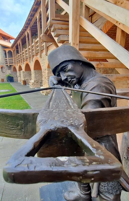 A bronze statue of an artisan wearing a cap is shown looking intently at a large anvil, set within a courtyard featuring wooden arcades and stone architecture.