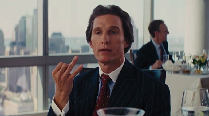 A still from a film showing a man in a dark pinstripe suit and red patterned tie, gesturing with his hand while seated at a restaurant table with a city skyline visible through the windows behind him.