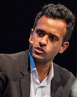 A man with short dark hair wearing a black suit jacket over a white shirt speaks into a small yellow headset microphone against a dark background.