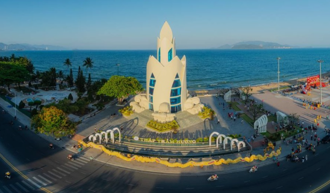 An aerial view of a coastal city featuring a tall, distinctive white monument shaped like a lotus or flame, surrounded by a roundabout and the ocean.